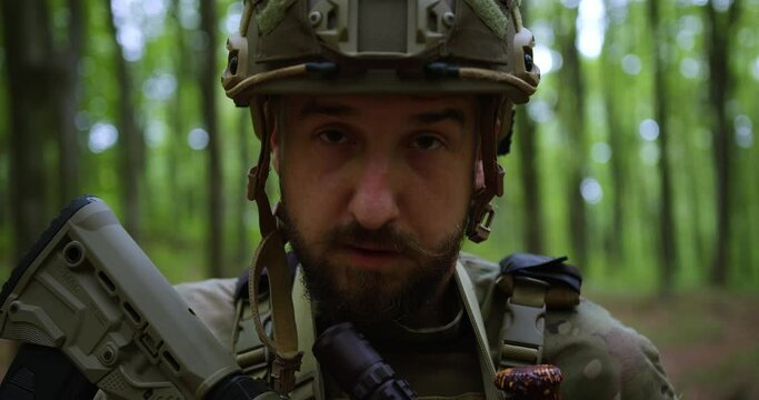 The evil face of the Ukrainian militant of the army soldier with weapons in his hands and a helmet on his head stands in the forest. A look at the camera. Sweat on the face. close-up