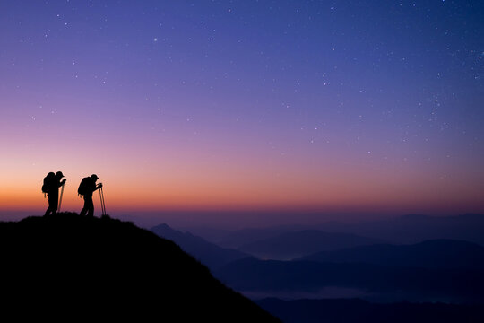 Silhouette of two young hikers were standing at the top of the mountain looking at the stars and Milky Way over the twilight sky. Both of them were happy and free to travel.