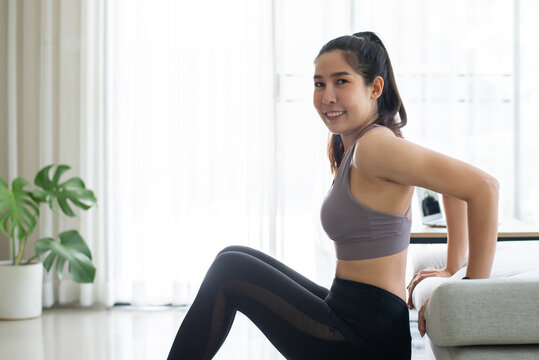 Asian young woman doing exercises with triceps dips poses to strengthen the upper arm muscles on the sofa in the living room at home. She likes to exercise while at home before going to work.
