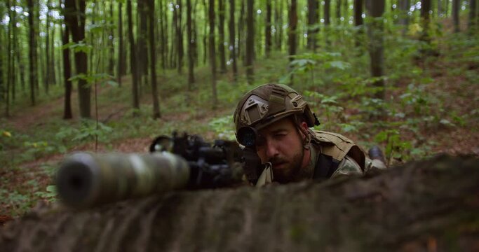 the sniper lies on the ground in his coat and aims at the optical sight with one eye closed. bearded soldier aims with weapons in the forest. switching focus from soldier to weapon silencer