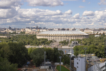 Naklejka premium Luzhniki sports stadium, green area with trees and bushes, building constructions and cable car across the river. Cityscape of Moscow. Cloudy sky on a summer day.