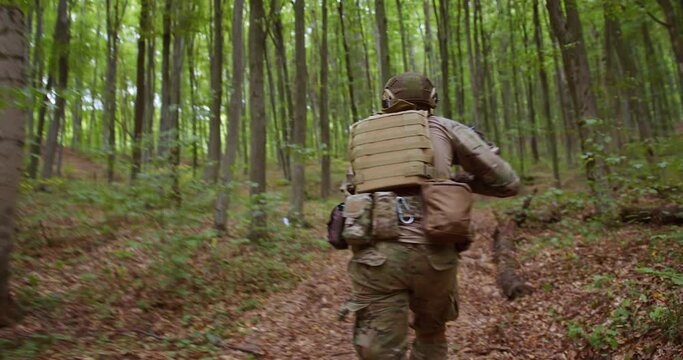 a soldier in ammunition, a bulletproof vest, a helmet and with a weapon in his hands runs through a large forest with tall trees. back view. dynamic camera. war in Ukraine. Ukrainian soldier