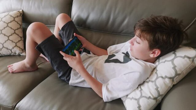 Happy boy playing emotionally game on smartphone is lying on sofa. Child plays bedtime. Internet. Gamer. Quarantine. Distance learning. Online education. Lifestyle. Hobby. Relax. Childhood