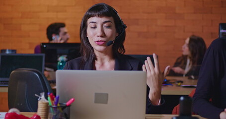 Confident young woman speaking with headset in front of computer screen at office call center. Female employee working at customer help desk talking