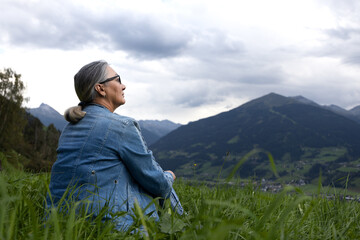 An elderly woman in a denim suit sits on an alpine meadow and looks towards the mountains.