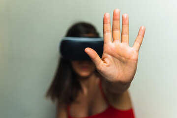 Woman with virtual reality glasses on neutral background seen from the front raising a hand making the stop gesture. Young girl in her room trying augmented reality in three dimensions.