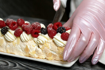 The woman cuts the cake. Hands in rubber gloves. Meringue roll with mascarpone cream. Garnished with raspberries and almond petals. Close-up.