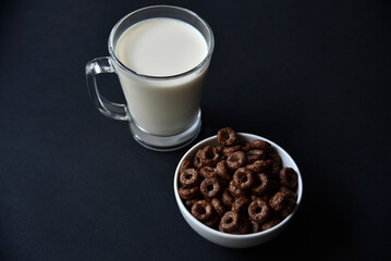 A glass of milk on a black background and chocolate corn rings. Delicious breakfast of cornflakes and milk.