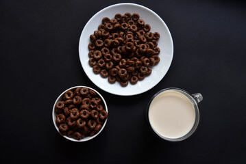 A saucer and a cup of cornflakes and a glass of milk on a black background. Delicious breakfast of cereal with honey and chocolate with milk.