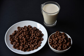 A saucer and a cup of cornflakes and a glass of milk on a black background. Delicious breakfast of cereal with honey and chocolate with milk.