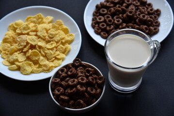 A saucer and a cup of cornflakes and a glass of milk on a black background. Delicious breakfast of cereal with honey and chocolate with milk.