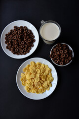 Obraz na płótnie Canvas A saucer and a cup of cornflakes and a glass of milk on a black background. Delicious breakfast of cereal with honey and chocolate with milk.