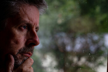 A man looks out from a wet rainy window. A distant thoughtful look on his face. He holds a pair of...
