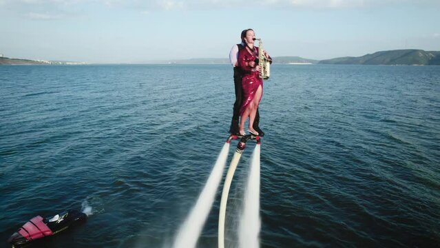 Show over the water on a flyboard. A woman in a red dress plays the saxophone, a man flies with her together.
