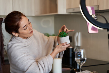 young happy influencer woman sitting in the dining room and having a video call with her phone, showing a succulent plant for sale to her followers