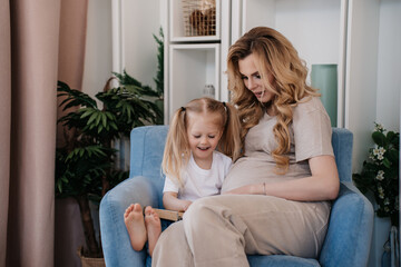 Pregnant swedish woman in beige t-shirt and pants sitting with her daughter in light blue chair, reading a book and wide smiling. Mom and daughter with tails at home, Maternity and pregnancy concept.
