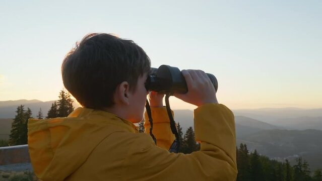 Happy Boy Looks Through Big Binoculars Into Distance Of Mountain Peaks Lens Flares In Autumn At Sunset On Sunny Day. Smily Happy Boy Hiker. Travel Concept. Tourism. Lifestyle. Dreamer. Go Everywhere 