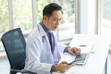 Portrait of asian senior doctor sitting in medical office while using laptop computer and checking time of appointment.