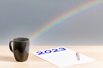 Number 2023 written on a paper, with a mug, a pen and a rainbow background. New year