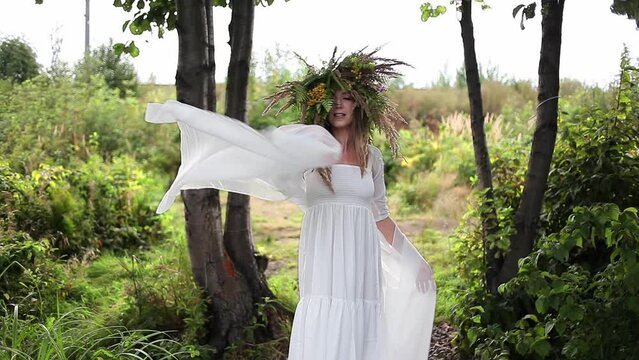 A young woman in a white dress and wearing a large wreath of field herbs dances in a meadow, unity with nature, an image in folk style