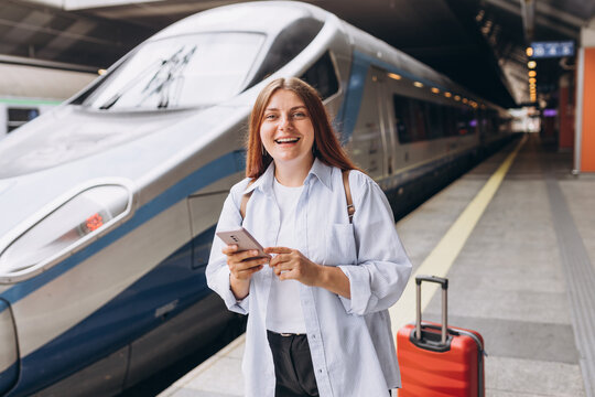 Young redhhead woman waiting train with backpack and using smart phone. Railroad transport concept, Traveler.Woman with suitcase walking at railroad station platform. Travel to vacation by train