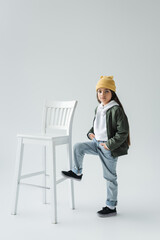 full length of cute girl in trendy autumnal outfit and beanie hat standing with hands in pockets near high chair on grey.