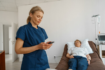 Nurse in uniform using phone standing near sick patient in hospital ward. High quality photo