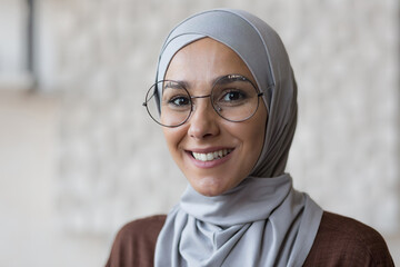 Close up photo portrait of young smiling Muslim woman in glasses and gray hijab, Arab woman at home smiling and looking at camera.