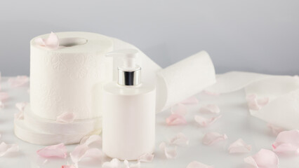 Fototapeta na wymiar Roll of toilet paper with cosmetic dispenser bottle on light background with rose petals and copy space. White toilet tissue, Hygiene product. Restroom soft touch toilet paper. Soft focus style image