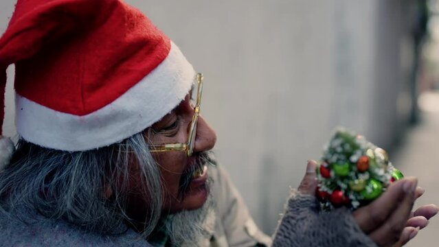 4k, An old homeless Asian man wearing a red Christmas hat holding a small Christmas tree is showing signs of a mental breakdown. The madman sat alone on the roadside laughing and smiling.