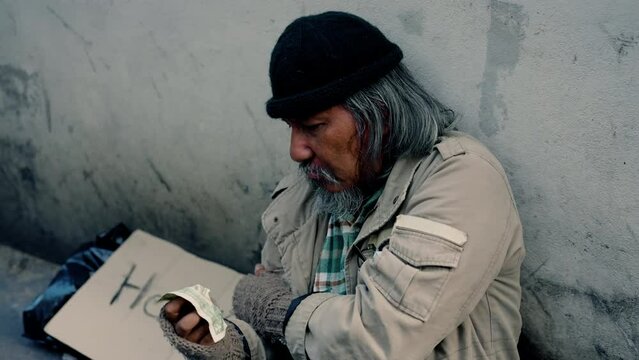 4k, An elderly Asian homeless man leaning against a wall with a sign for help next to it when someone brings him a banknote refuses to accept it because he needs more work. Homeless not have home