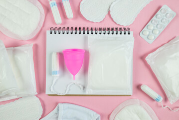 Top view photo of open planner over sanitary napkins, tampons, underwear and pills on isolated pastel pink background