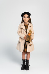 full length of cute girl in stylish trench coat and beret standing with toy horse on grey.