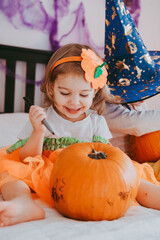 Little toddler girl drawing on a pumpkin making lantern jack on Halloween holiday. Little girl in a carnival costume celebrating halloween at home