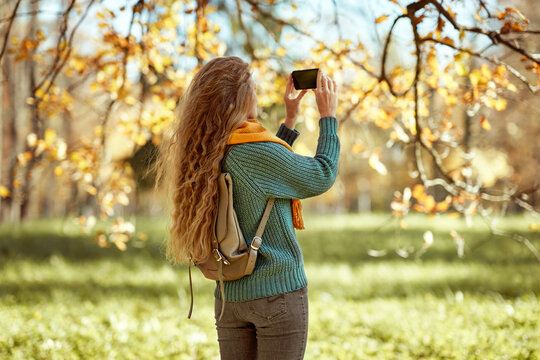 A young red-haired woman walks through the autumn park. A girl with a smartphone takes photos in an autumn forest