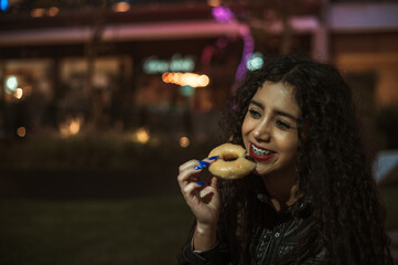 Brunette girl with curly hair enjoys a donut in a place full of lights at nightfall, she wears a...