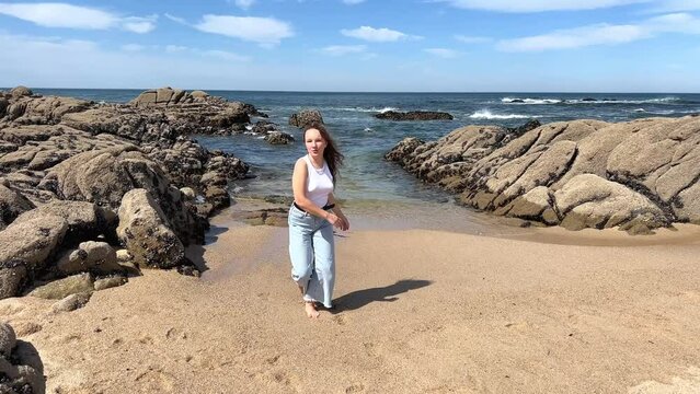 a teenage girl with blond hair in a white T-shirt and jeans is spinning against the backdrop of the Atlantic Ocean in Portugal she is happy then she jumps joyfully smiling the tour was a success 4k