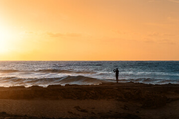 Fototapeta na wymiar Mann steht am Strand Spiaggia di Scivu auf Sardinien Italien und fotografiert den Sonnenuntergang