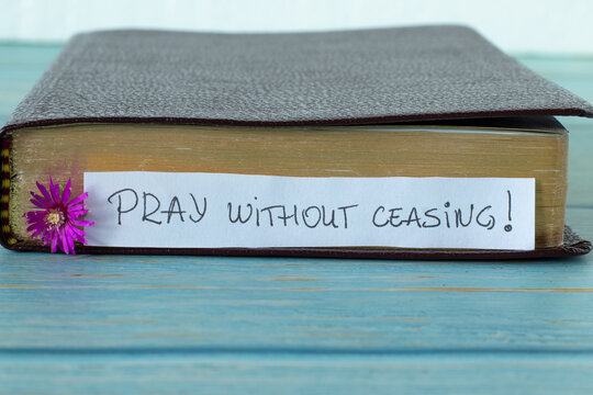 Pray without ceasing, a handwritten text on a note with a purple flower in front of a closed Holy Bible Book with golden pages placed on a blue wooden table with white background. A close-up.