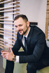 A young man with a beard, dressed in a classic business suit, sitting in the hands of a glass of whiskey. Portrait of a man relaxing with a glass of hard liquor.