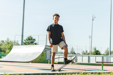 Afro-American boy with black t-shirt posing with his skateboard with the sky in the background