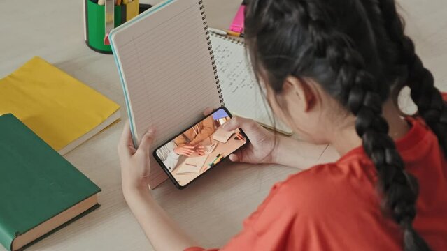 Over the shoulder shot of school girl sitting at desk, hiding smartphone behind copybook and watching video on the Internet during class time
