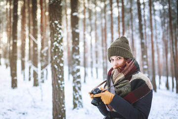 Fototapeta na wymiar Winter scenery. A woman in a winter hat and a scarf holds a camera in her hands. In the background, forest trees in the snow.