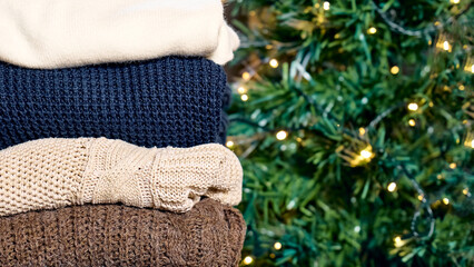 Stack of warm knitted sweaters on blurred christmas tree background. Slow Fashion. Conscious consumption. Black friday. Winter sale.