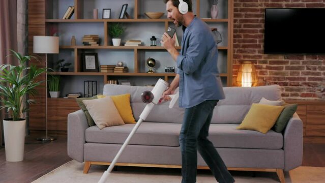 Cheerful guy, the man cleans the apartment. Vacuuming, wearing headphones with dancing and singing into a smartphone instead of a microphone. House cleaning is easy.