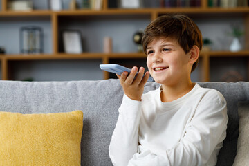 Young cute boy recording voice message on smartphone. Smiling teenager using virtual assistant app...