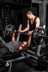 High quality photography. Latino man in a black gym lifting 90 pounds while his friend helps him.