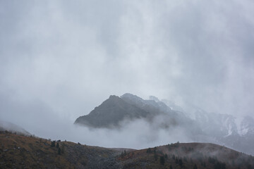 Dark atmospheric landscape with high mountain silhouettes in dense fog in rainy weather. Snowy rocky mountain top above hills in thick fog in dramatic overcast. Black rocks in low clouds during rain.