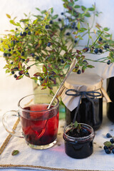 On a light background, on a beautiful fabric napkin, there are several packaged glass jars with blueberry jam, and in front there is a fresh tasty blueberry with green leaves on the branches and a swe
