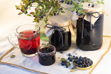 On a light background, on a beautiful fabric napkin, there are several packaged glass jars with blueberry jam, and in front there is a fresh tasty blueberry with green leaves on the branches and a swe